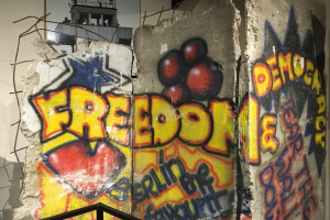 Graffiti mit der Aufschrift "Freiheit und Demokratie" in schwarzer Großbuchstaben-Schrift auf einer Wand, mit einem Geländer unten und einem Gebäude mit Fenstern im Hintergrund.