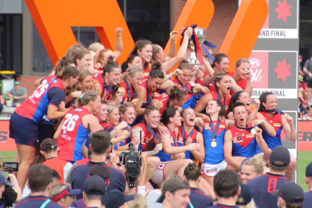 Eine Gruppe von Frauen-AFL-Spielerinnen feiert mit einem Pokal vor einer jubelnden Menge, einige tragen Medaillen und Kappen, andere halten Kameras, mit Tafeln und einem Gebäude im Hintergrund.