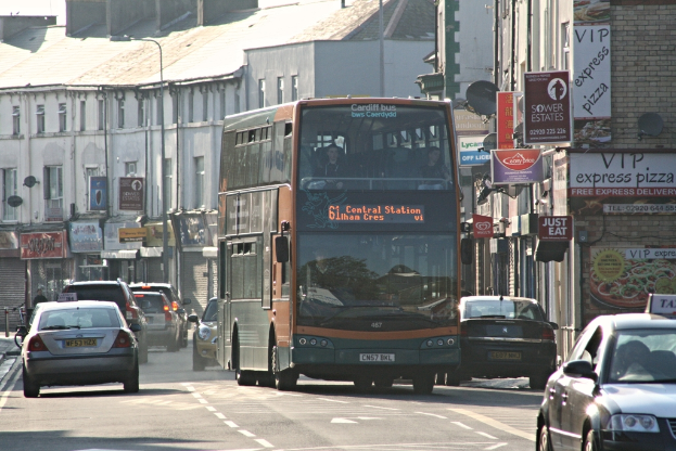 Eine Straße mit Autos und einem Bus vor Gebäuden mit Wänden, Fenstern, Tellern und Dächern, mit Plakaten und Bannern an den Wänden und einem Mast mit Straßenlaterne.