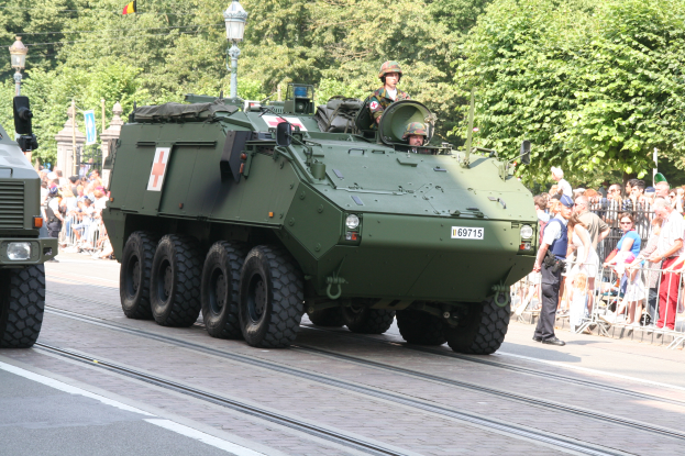 Ein militärisches Fahrzeug fährt auf einer Straße mit Geländern an einer Menschenmenge vorbei, neben der einige Helme tragen, mit Bäumen, Laternen, Fahnen und einem klaren blauen Himmel im Hintergrund.