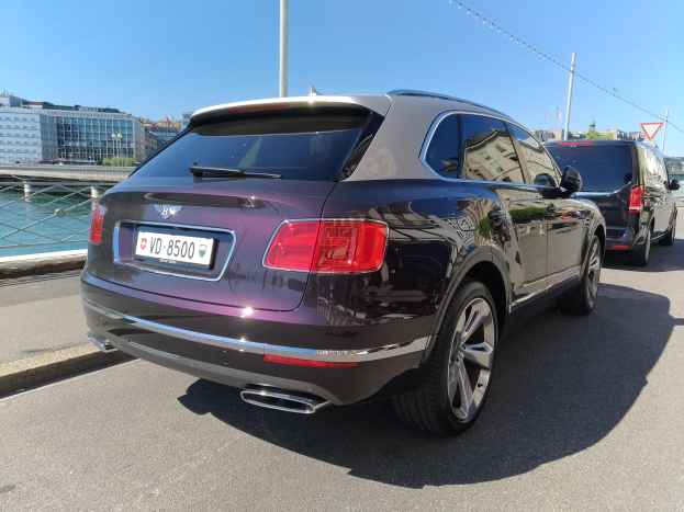 Ein Bentley Bentayga V8 S fährt auf einer Straße mit einer Brücke, Gebäuden, Bäumen und einem klaren blauen Himmel im Hintergrund.