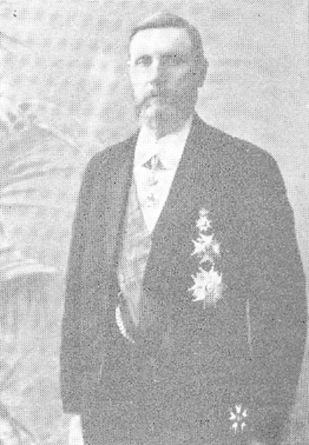 Ein Mann in einem schwarzen und weißen Anzug mit einem weißen Hemd und schwarzer Krawatte steht vor einer üppigen grünen Pflanze und schaut ernst.