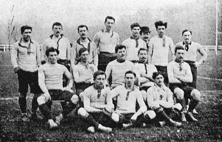 Schwarzes und weißes Foto einer Fußballmannschaft aus den 1920er Jahren, einige stehen, andere sitzen auf dem Boden, mit Pfählen, Bäumen und einem klaren Himmel im Hintergrund.