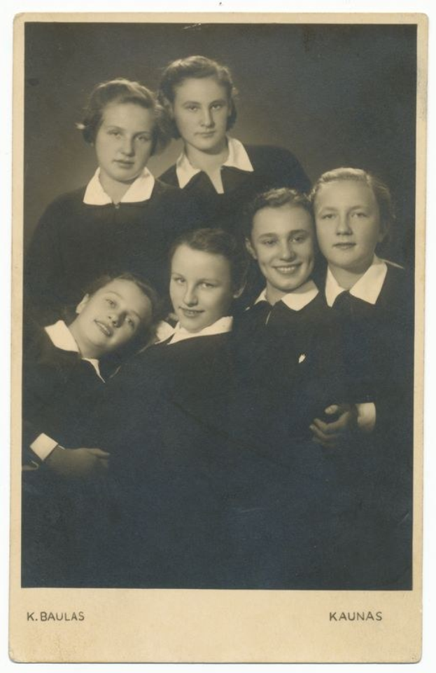 Ein Schwarz-Weiß-Foto einer Gruppe junger Mädchen in Schuluniformen, die fröhlich posieren und in die Kamera lächeln, mit Text unten.