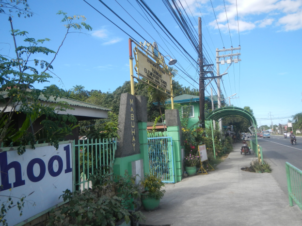 Schuleingangsschild am Straßenrand mit einem Bogen, Tor und Namenskennzeichen, umgeben von Fahrzeugen, Pflanzen, Bäumen, Strommasten mit Drähten und einem bewölkten Himmel.