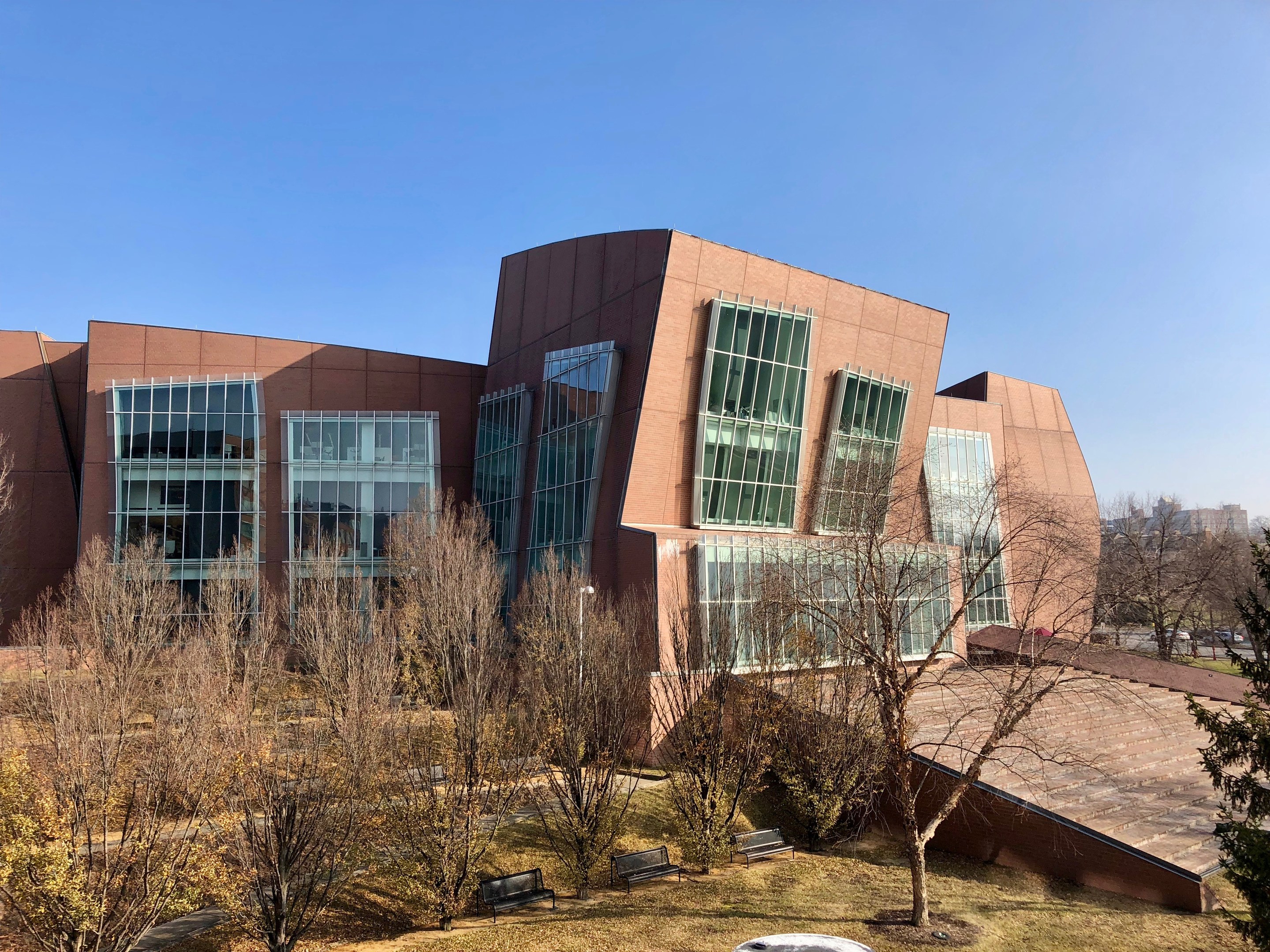 Großes modernes Gebäude mit zahlreichen Fenstern, umgeben von Bäumen, Bänken, Gras und einem Lichtmast, mit anderen Gebäuden und einem klaren blauen Himmel im Hintergrund, als der neue Campus der University of Illinois at Chicago identifiziert.