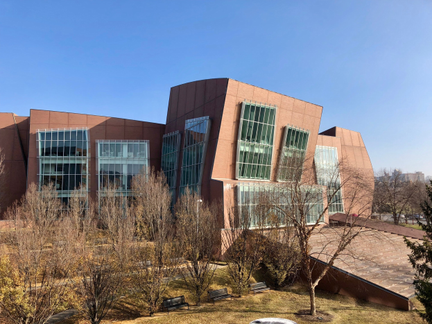 Großes modernes Gebäude mit zahlreichen Fenstern, umgeben von Bäumen, Bänken, Gras und einem Lichtmast, mit anderen Gebäuden und einem klaren blauen Himmel im Hintergrund, als der neue Campus der University of Illinois at Chicago identifiziert.