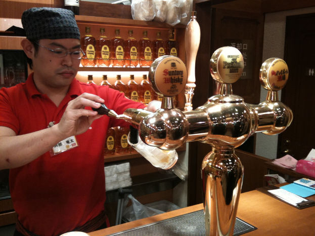 Eine Person mit Mütze und Brille füllt ein Glas mit Bier an einem Tisch mit Papieren und einer Maschine, mit Regalen voller Alkohol im Hintergrund.