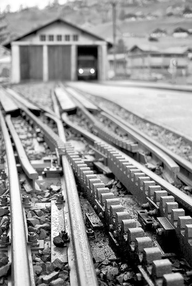 Eisenbahnschienen verlaufen durch eine Szene mit einer Hütte, einem Fahrzeug und Häusern im Hintergrund.