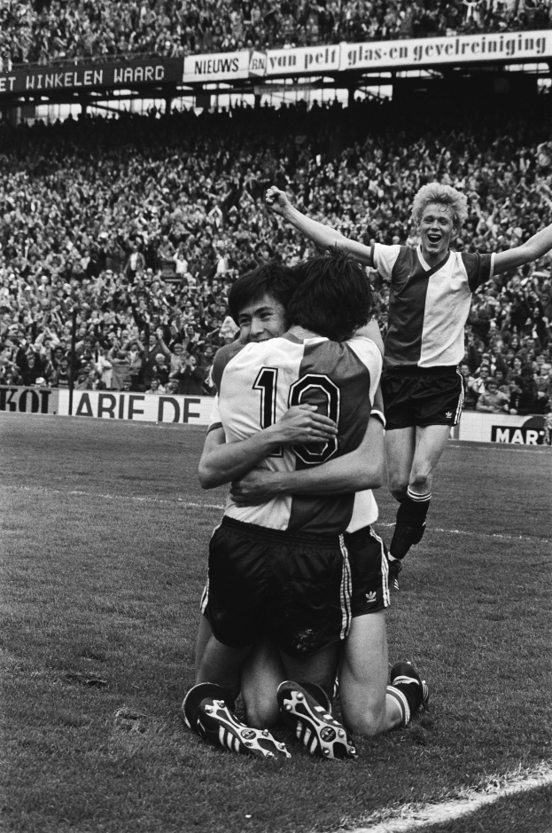 Drei Fußballspieler, die sich nach einem Tor auf dem Boden umarmen, mit einer jubelnden Menge und schwenkenden Fahnen in einem schwarz-weißen Stadion Setting.