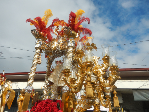 Ein großer goldener und roter Festwagen, geschmückt mit Blumen und anderen Dekorationen bei einem Karnevalsumzug, mit einem Gebäude, Strommasten mit Drähten und einem bewölkten Himmel im Hintergrund.