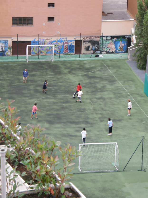 Kinder beim Fußballspielen auf einem Feld mit Toren, ein Gebäude mit Graffiti im Hintergrund, Pflanzen links und Bäume rechts.