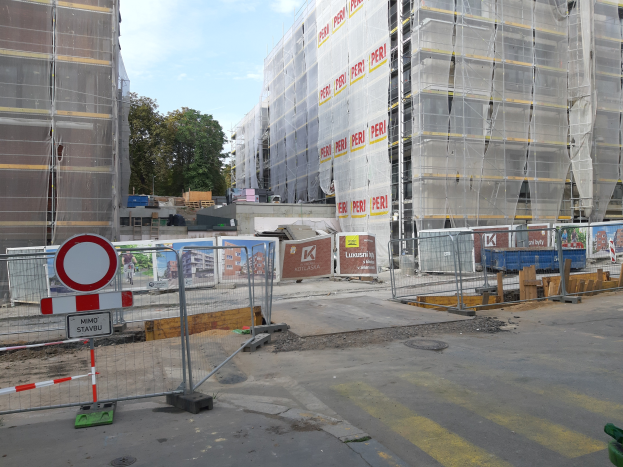 Eine Baustelle mit einem teilweise errichteten Gebäude im Hintergrund, umgeben von Bäumen unter einem klaren blauen Himmel, mit einer Straße auf der rechten Seite, die Absperrungen und ein Schild mit laufenden Bauarbeiten zeigt.