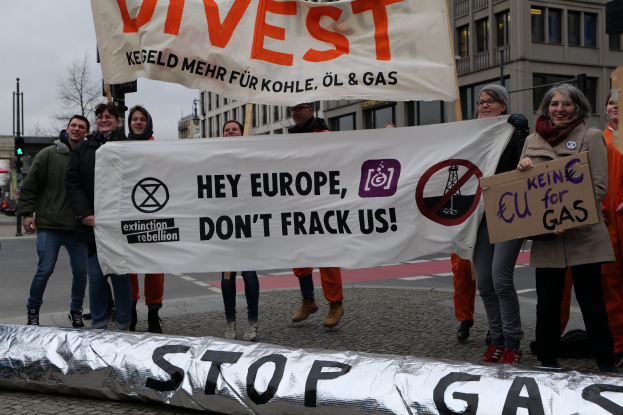 Gruppe von Menschen mit einem 'Stoppt Gas'Transparent auf der Straße bei einer Protestaktion gegen die Europäische Union vor dem Hintergrund von Gebäuden, Bäumen, Masten, Ampeln und einem klaren blauen Himmel.