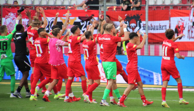 Gruppe von Fußballspielern, die ein Tor feiern, mit Flaschen oder klatschenden Händen, sowie Bannern, einem Zaun, Metallstangen, Stühlen und einer Decke mit Lampen im Hintergrund.