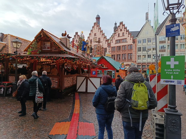 Menschen auf Kopfsteinpflasterstraße neben einem Weihnachtsmarkt in Nürnberg, Deutschland, mit Leuchten und Texttafeln im Vordergrund und Gebäuden im Hintergrund unter einem bewölktem Himmel.