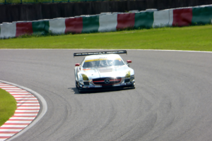 Ein Mercedes-Benz SLS AMG GT3 ist auf einer Straße am Nürburgring positioniert, umgeben von grünem Gras und einem Zaun im Hintergrund.