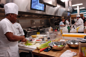Eine Gruppe von Köchen in einer Küche, die Essen auf einem Schneidbrett zubereiten, mit einem Koch im Vordergrund in einer weißen Schürze und Mütze, der ein Messer hält und von verschiedenen Utensilien, Schüsseln, Gemüse, einem Handy, Flaschen und anderen Gegenständen umgeben ist, während im Hintergrund ein Fernseher zu sehen ist.