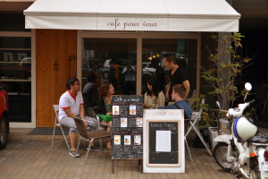 Ein weißes Haus mit einer Fahne, einem Motorrad, Topfpflanzen und mehreren Stühlen, mit einer Gruppe von Menschen in der Nähe, die sich in Autos und Lichtern spiegelt.
