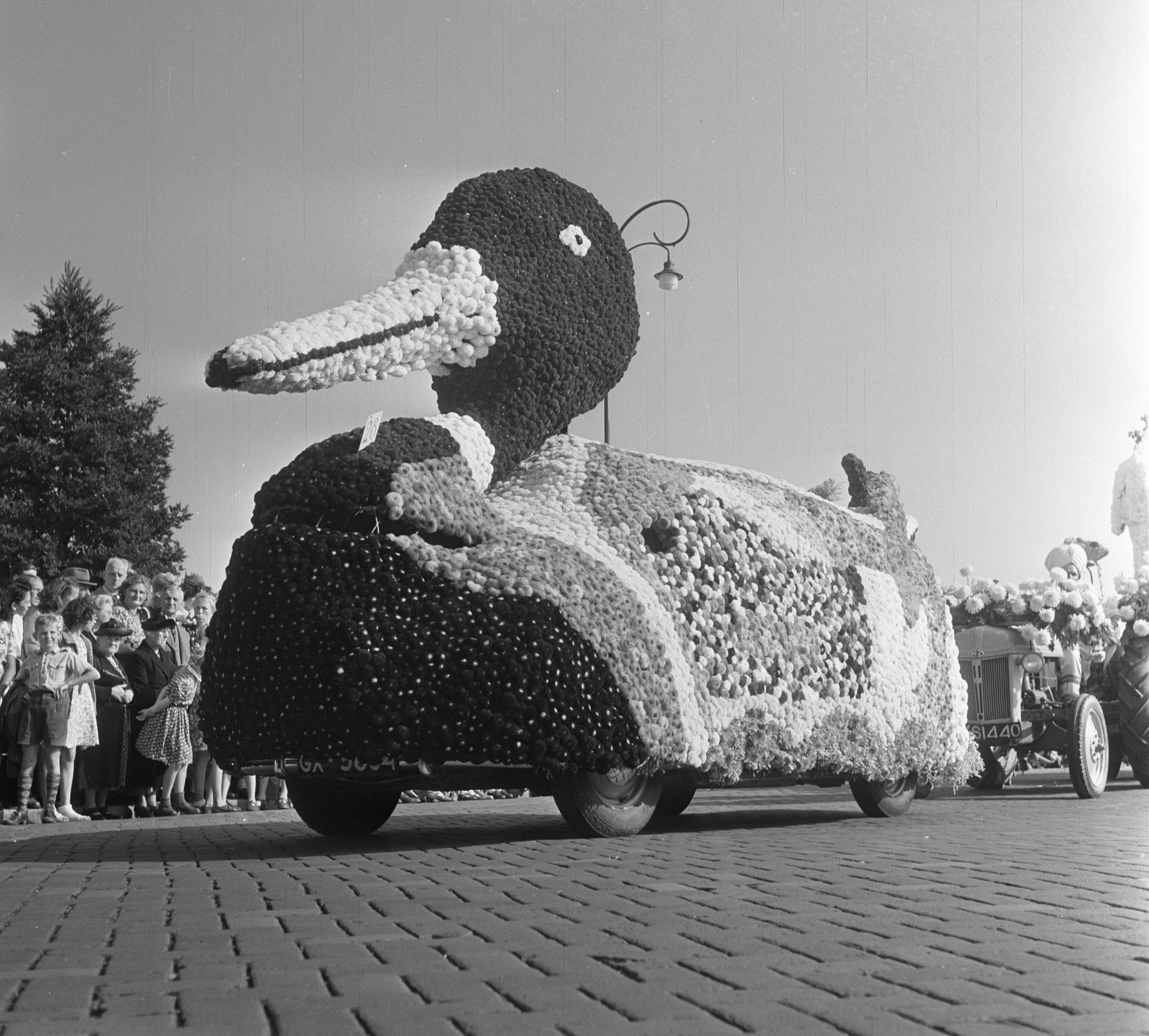 Schwarz-Weiß-Foto eines Paradewagens in Form einer Ente, dekoriert mit Blumen, mit Menschen links und Bäumen im Hintergrund.