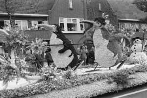 Schwarzes und weißes Foto eines Paradewagens, der mit Blumen und Pinguinfiguren geschmückt ist, mit Zuschauern, Bäumen und Gebäuden im Hintergrund.