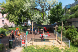 Ein Spielplatz mit spielenden Kindern, umgeben von einem Metallzaun, Gras, Pflanzen, einem Mülleimer, einem Schild an einem Pfahl, einigen Pfählen mit daran gebundenen Tüchern, Fahrzeugen, Gebäuden mit Fenstern und einem Baum im Hintergrund unter einem bewölkten Himmel.