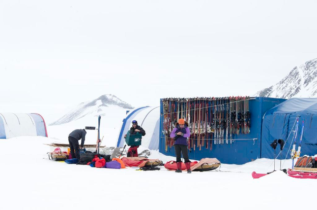 Drei Personen stehen auf einem schneebedeckten Eisland mit verstreuten Taschen, Zelten mit Skiern im Hintergrund und schneebedeckten Hügeln unter einem klaren Himmel.