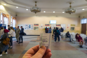 Eine Person hält ein Ticket vor einer Menge hoch, einige sitzen, andere stehen, ein Mann und eine Frau in der Nähe und eine Wand mit Postern und einem Bildschirm im Hintergrund, unter einer Decke mit Lampen und Kronleuchtern.