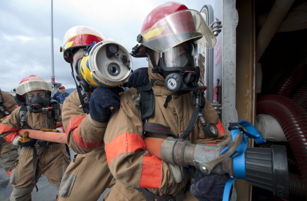 Feuerwehrleute in Schutzausrüstung mit einem Schlauch haltenden, vor einer Stange und Rohren gegen einen bewölkten Himmel stehend.