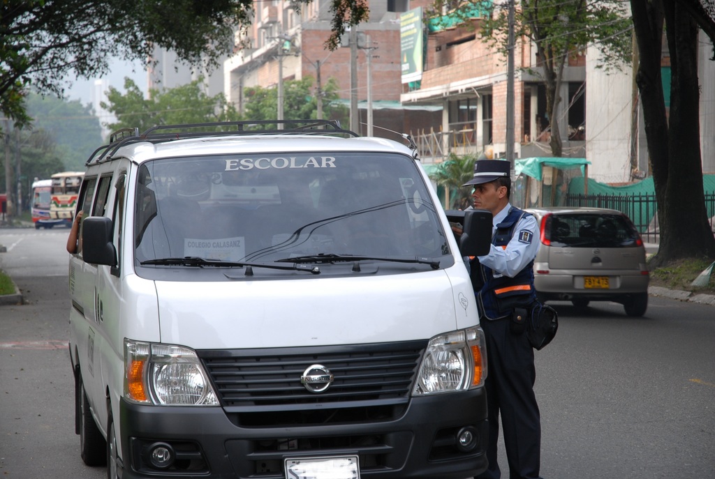 Ein Polizeibeamter spricht mit einer Person in einem Fahrzeug auf einer belebten Straße, umgeben von Bäumen, hellbraunen Gebäuden und Strommasten.