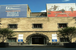 Außenansicht des Deutschen Sport & Olympia Museums in Berlin mit Fenstern, einer Tür, Bäumen und Bannern vor einem bewölkten Himmel.