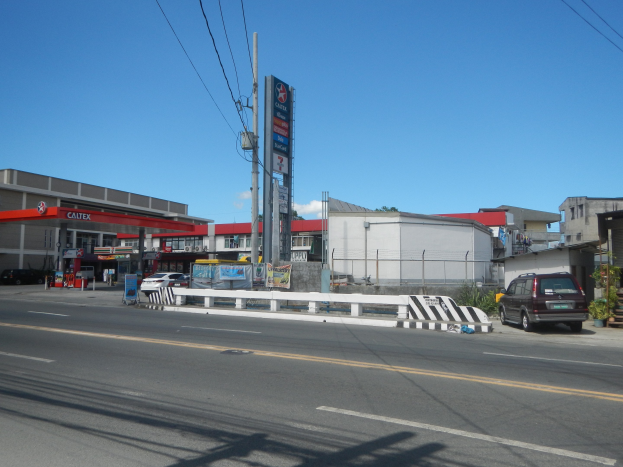 Tankstelle an einer Straßenecke mit parkenden Autos, Gebäuden, Strommasten, Schildern, Geländern, Pflanzen und einem bewölkten Himmel.