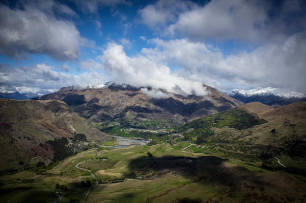 Eine Panoramansicht vom Gipfel eines Bergs in Queenstown, Neuseeland, zeigt saftiges grünes Gras, verstreute Bäume, eine gewundene Straße und einen Himmel voller weißer, flauschiger Wolken.