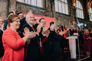 Eine Gruppe von Menschen, wahrscheinlich Liberale, klatscht vor einer Menge, mit einem Podium, Mikrofon und Texttafel rechts, Stühlen, einem Banner, einer Wand mit Fenstern und Lichtern im Hintergrund.