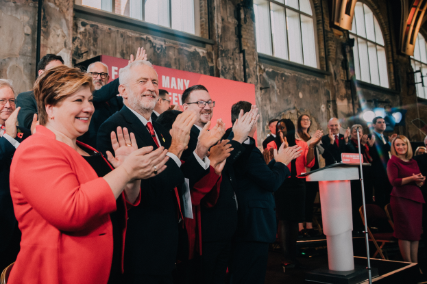 Eine Gruppe von Menschen, wahrscheinlich Liberale, klatscht vor einer Menge, mit einem Podium, Mikrofon und Texttafel rechts, Stühlen, einem Banner, einer Wand mit Fenstern und Lichtern im Hintergrund.