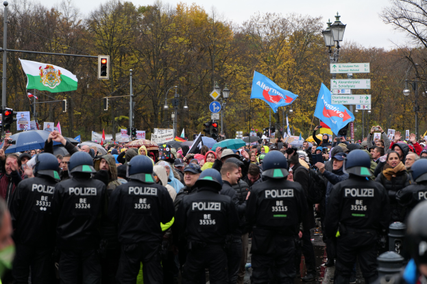 Eine gro├če Gruppe von Polizisten steht vor einer Menge mit Fahnen und Schirmen, mit Laternenmasten, Verkehrsampeln, Verkehrszeichen, B├Ąumen und einem klaren Himmel im Hintergrund.