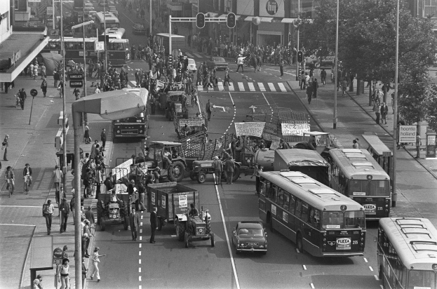 Schwarzes und weißes Foto einer belebten Stadtstraße mit Bussen, Autos, Lastwagen, Fußgängern auf dem Gehweg, Ampeln, Strommasten, Bäumen und Gebäuden im Hintergrund.