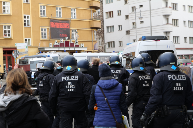 Polizeibeamte in Uniform vor einer Menge bei einer Demonstration in Berlin, mit Fahrzeugen, Gebäuden und einer Kamera im Bild.