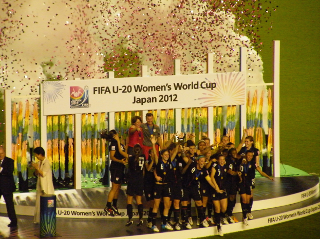 Frauenfußballspielerinnen in schwarzen Trikots feiern auf einer Bühne mit Publikum und dekoriertem Hintergrund.