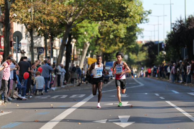 Zwei Männer bei einem Marathon auf einer Stadtstraße mit einer Menge auf der linken Seite und im Hintergrund Gebäude, Bäume und einen klaren Himmel.
