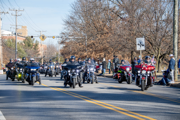 Eine Gruppe von Menschen auf Motorrädern, die eine Straße entlangfahren, die von Strommasten, Schildern, Bäumen und Gebäuden gesäumt ist, unter einem klaren blauen Himmel, einige tragen Helme.