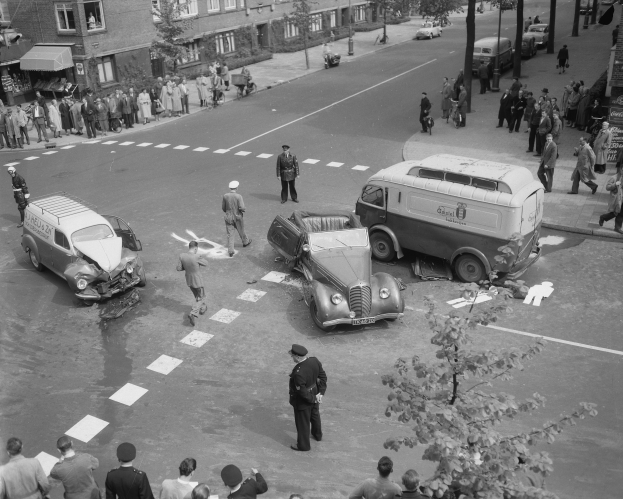 Eine Schwarz-Weiß-Szene eines Autounfalls an einer Straßenkreuzung mit mehreren Fahrzeugen, darunter ein Lieferwagen, einer Gruppe von Menschen drumherum und im Hintergrund Gebäude, Bäume, Pfosten und Texttafeln.
