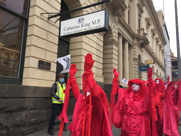 Eine Gruppe von Menschen in roten Kostümen geht während eines Ballarat-Faschingsumzugs eine Straße entlang, mit einer Person in einer grünen Jacke und einem Hut auf der linken Seite, mehreren Brettern, die an einem Pfahl befestigt sind, vor einem Gebäude und dem Himmel oben im Bild.