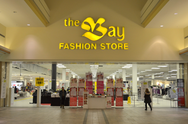 Außenansicht eines Bay Fashion Stores in einem Einkaufszentrum mit Menschen, Texttafeln an Wänden, Säulen, Deckenleuchten und Hintergrunddisplays.