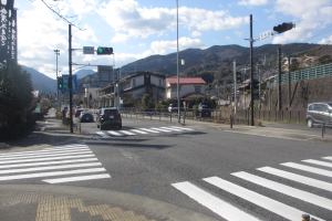 Eine Stadtstraße mit Autos, einer Fußgängerüberführung, Strommasten mit Leitungen, Verkehrsampeln, Gebäuden mit Fenstern, Bäumen, Hügeln und einem bewölkten Himmel im Hintergrund.