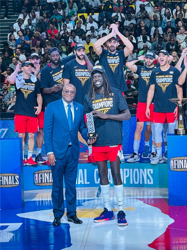 Eine Gruppe von Männern auf einem Basketballfeld feiert mit Trophäen, während andere im Hintergrund sitzen und zwei Trophäen rechts auf Podesten stehen.