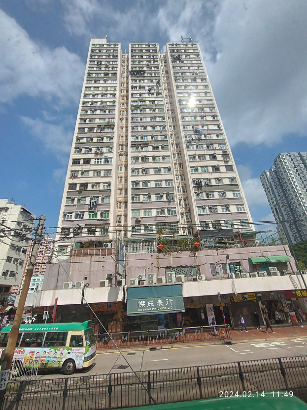 Ein hoher Wohnblock auf einer belebten Stadtstraße umgeben von anderen Gebäuden, Fahrzeugen, Passanten und urbanen Elementen unter einem bewölkten Himmel.