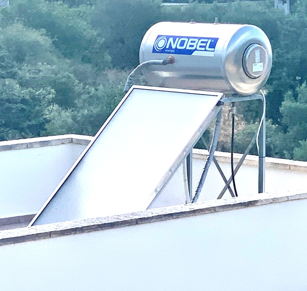 Solarwassererwärmer mit einem Speicherbehälter auf dem Dach eines Gebäudes, umgeben von Bäumen im Hintergrund.