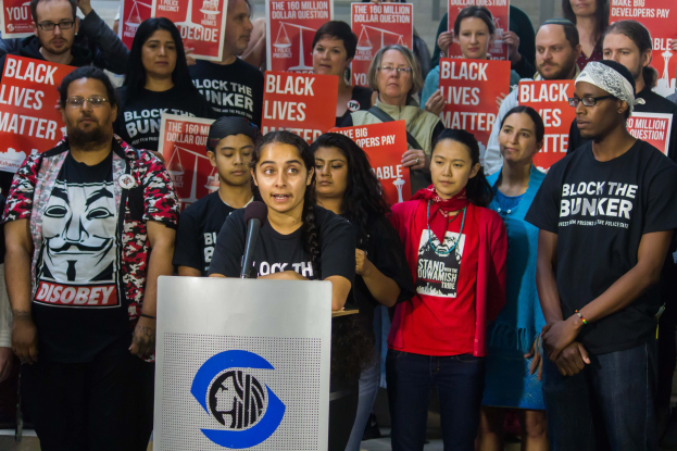 Eine Gruppe von Menschen mit "Black Lives Matter"-Plakaten, mit einer Frau an einem Rednerpult mit Mikrofon in der Mitte, die vor einer Menge und einer Wand steht.