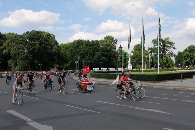 Eine Gruppe von Menschen, die auf Fahrrädern eine von Bäumen gesäumte Straße mit Laternenmasten und Fahnenmasten entlangfahren, bei bewölktem Himmel und fahrenden Fahrzeugen im Hintergrund.